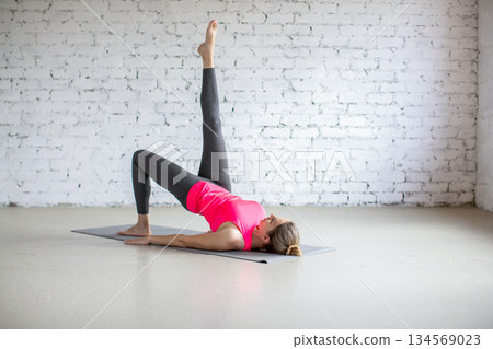 Slim Caucasian 40 years old woman lying on back and does shoulder bridge exercise with one leg up in loft studio indoor. Slim Caucasian 40 years old woman lying on back and does shoulder bridge exercise with one leg up in loft studio indoor. 134569023