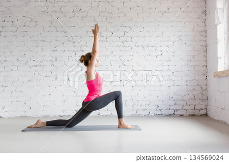Slim Caucasian woman practice Ashva Sanchalanasana exercise with rising hands in loft fitness studio indoor. Slim Caucasian woman practice Ashva Sanchalanasana exercise with rising hands in loft fitness studio indoor. 134569024