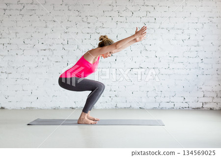 Caucasian slim woman in pink sportswear practice chair pose indoor in loft fitness studio, Utkatasana. Caucasian slim woman in pink sportswear practice chair pose indoor in loft fitness studio, Utkatasana. 134569025