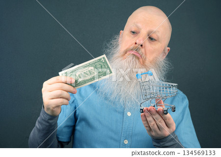 bald man with a large white beard holds a dollar and a model of a grocery basket 134569133