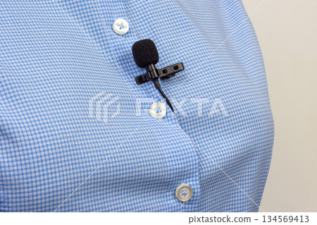 A compact black lav mic is secured on a white buttoned blue shirt A compact black lav mic is secured on a white buttoned blue shirt 134569413