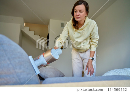 Woman using handheld vacuum cleaner to clean sofa in modern living room Woman using handheld vacuum cleaner to clean sofa in modern living room 134569532