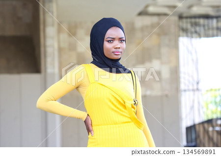 Confident African woman in yellow dress and hijab posing outdoors during daylight 134569891