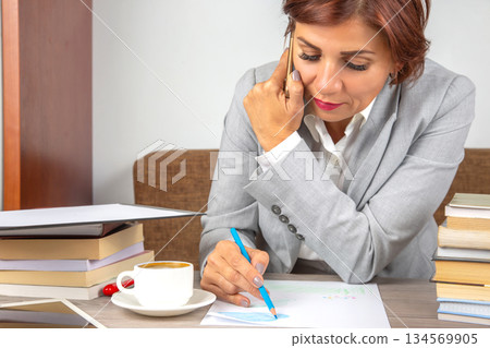Woman in gray suit studies documents while writing notes at modern office desk Woman in gray suit studies documents while writing notes at modern office desk 134569905