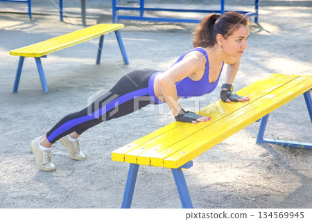 Woman performs push-ups on yellow wooden benches in an outdoor fitness area during daytime 134569945