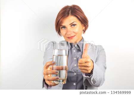 Woman in gray suit holds glass of water and gives thumbs up gesture with a smile Woman in gray suit holds glass of water and gives thumbs up gesture with a smile 134569999