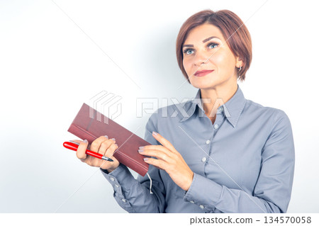 Woman in gray shirt holding a notebook and pen while looking thoughtfully at the ceiling 134570058