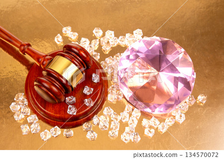 Gavel placed next to a large pink gemstone and sparkling diamonds on a golden background 134570072
