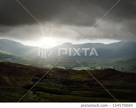 Sunlight streams through a break in the clouds, illuminating a valley within the Coomloughra Horseshoe mountains. A hiking trail winds through the landscape in Ireland. Sunlight streams through a break in the clouds, illuminating a valley within the Coomloughra Horseshoe mountains. A hiking trail winds through the landscape in Ireland. 134570084