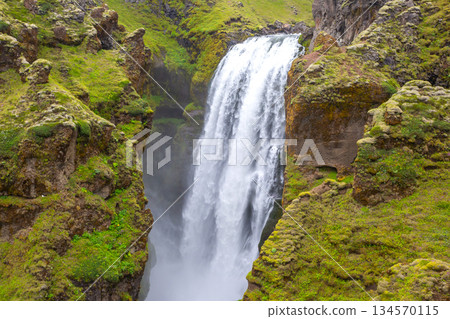 Majestic waterfall cascading over lush green cliffs in Iceland's pristine landscape 134570115