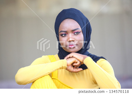Young african Woman in a Black Hijab and Yellow Dress Reflecting Thoughtfully in an Outdoor Setting 134570118