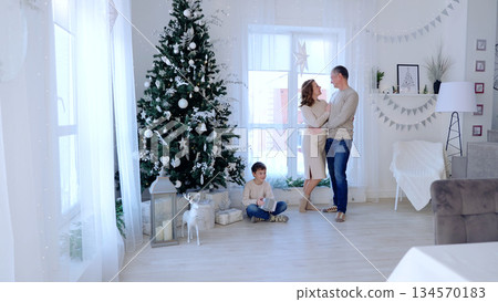 Happy family enjoying the holiday season near a decorated christmas tree in their cozy living room, with the son holding a gift. A day with family before Christmas. New Year's Eve with family Happy family enjoying the holiday season near a decorated christmas tree in their cozy living room, with the son holding a gift. A day with family before Christmas. New Year's Eve with family 134570183