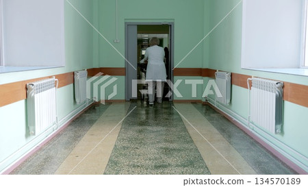 Russia Tomsk 05.07.2025: Surgical nurse escorting patient through corridor, pale mint walls, worn flooring, clinical lighting, measured pace during postoperative handover, focused staff 134570189