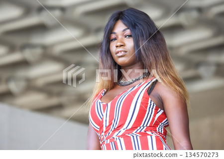 Confident African woman posing in stylish striped dress against modern architectural backdrop Confident African woman posing in stylish striped dress against modern architectural backdrop 134570273
