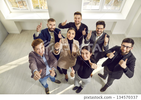 Team of happy business people standing in office, looking up, smiling and giving thumbs up Team of happy business people standing in office, looking up, smiling and giving thumbs up 134570337