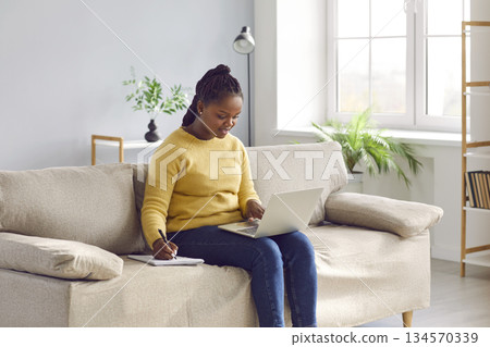 African American woman sitting on sofa, using laptop, studying online, and taking notes African American woman sitting on sofa, using laptop, studying online, and taking notes 134570339