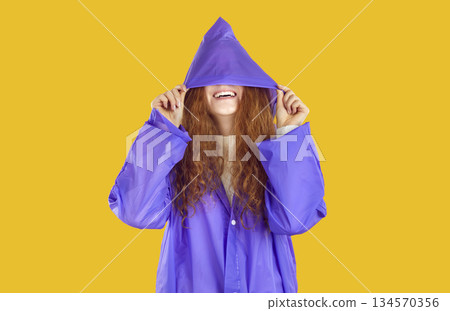 Smiling curly redhead girl in purple raincoat hiding in hood on her eyes on yellow background. 134570356