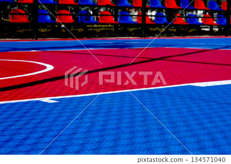 Vibrant basketball court with red and blue colors under bright sunlight, showing court markings and shadows during daytime activity Vibrant basketball court with red and blue colors under bright sunlight, showing court markings and shadows during daytime activity 134571040
