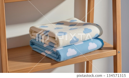 cloud-patterned baby blanket, cotton material, ivory and sky blue tones, folded neatly on a wooden shelf, morning sunlight, soft shadows 134571315
