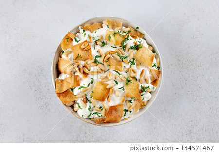 Levantine fatteh with pita chips, chickpeas, yogurt sauce, pine nuts and parsley on white background. Top view. Levantine fatteh with pita chips, chickpeas, yogurt sauce, pine nuts and parsley on white background. Top view. 134571467