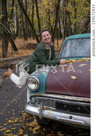 Smiling woman leans on a rusty vintage car with turquoise and brown paint, fallen yellow leaves, autumn forest in background 134571833