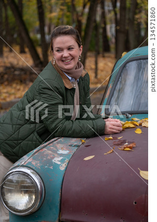 Smiling woman leans on a rusty vintage car with turquoise and brown paint, fallen yellow leaves, autumn forest in background Smiling woman leans on a rusty vintage car with turquoise and brown paint, fallen yellow leaves, autumn forest in background 134571860