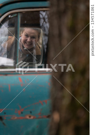 Smiling woman looks through the window of vintage turquoise car, peeling paint, soft autumn light, shallow depth of field, warm nostalgic mood 134571861