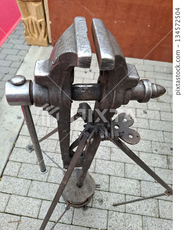 An antique vise stands on a sturdy base, ready for use in metal forging. This tool holds metal securely while craftsmen shape and work on projects. It is a key part of the workshop. 134572504