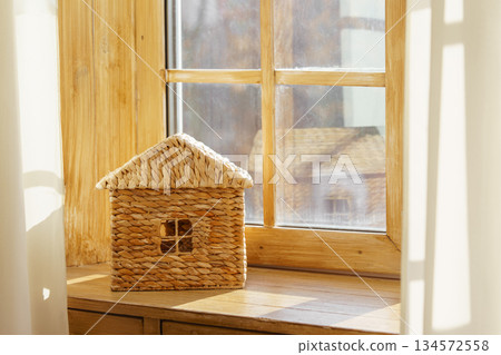 Small woven house model on sunny wooden window sill against natural light window in child room 134572558