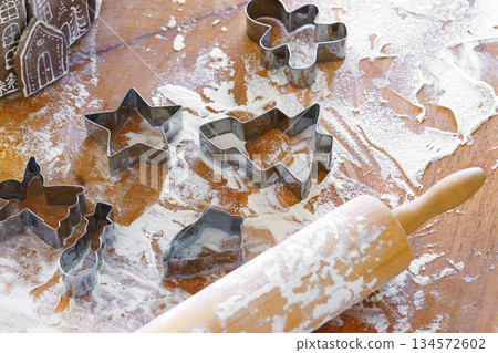 Baking gingerbread cookie cutters, wooden rolling pin, and flour are messily spread on kitchen table 134572602