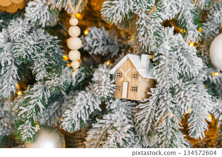 Snowy Christmas tree branch with wooden house ornament and warm golden holiday light illumination 134572604