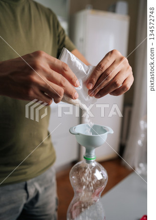 Vertical cropped shot of unrecognizable man gardener carefully pouring fertilizer from plastic bag into funnel inserted into bottle, preparing fertilizer solution for plants. Concept of indoor farming Vertical cropped shot of unrecognizable man gardener carefully pouring fertilizer from plastic bag into funnel inserted into bottle, preparing fertilizer solution for plants. Concept of indoor farming 134572748