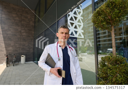 Healthcare professional stands outside modern clinic holding a tablet and phone in the daytime 134575317