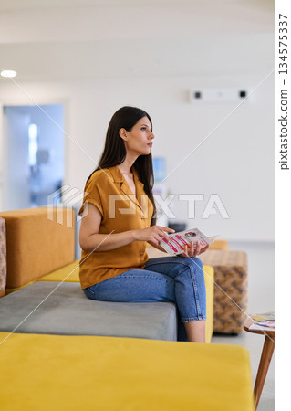 Woman sitting on a bench in a modern waiting area, engaged in reading a magazine while enjoying a quiet moment Woman sitting on a bench in a modern waiting area, engaged in reading a magazine while enjoying a quiet moment 134575337