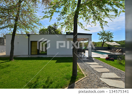 Covered terrace of a modern modular house with dining table, overlooking a swimming pool and gabion fence. Contemporary prefabricated home architecture with outdoor living space Covered terrace of a modern modular house with dining table, overlooking a swimming pool and gabion fence. Contemporary prefabricated home architecture with outdoor living space 134575341