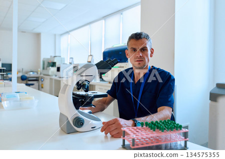 Lab technician using a microscope to analyze samples in a bright, modern laboratory environment during work hours Lab technician using a microscope to analyze samples in a bright, modern laboratory environment during work hours 134575355