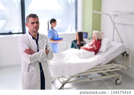 Doctor standing with arms crossed in hospital room while nurses attend to patient in bed Doctor standing with arms crossed in hospital room while nurses attend to patient in bed 134575419