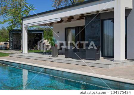 Modern modular house backyard featuring a rectangular swimming pool, large patio with wooden sun loungers, and umbrellas against a blue sky. Contemporary prefabricated home design Modern modular house backyard featuring a rectangular swimming pool, large patio with wooden sun loungers, and umbrellas against a blue sky. Contemporary prefabricated home design 134575424