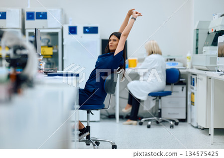 Lab technicians working in a modern laboratory preparing samples while one stretches during a busy workday 134575425