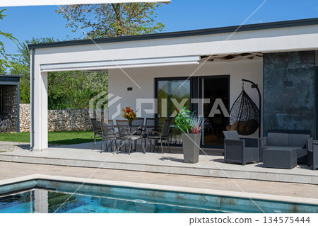 Modern modular house backyard featuring a rectangular swimming pool, large patio with wooden sun loungers, and umbrellas against a blue sky. Contemporary prefabricated home design 134575444