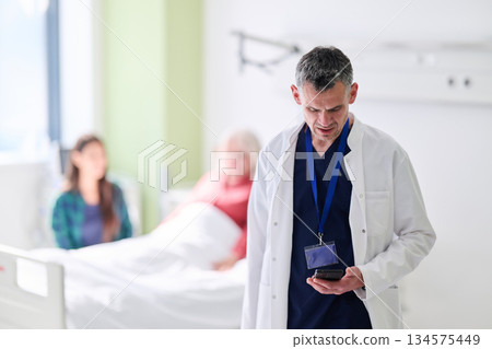 Doctor checks device while patient rests in hospital room during daytime consultation 134575449