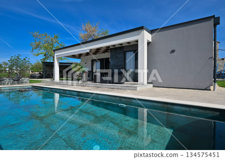 Modern modular house backyard featuring a rectangular swimming pool, large patio with wooden sun loungers, and umbrellas against a blue sky. Contemporary prefabricated home design Modern modular house backyard featuring a rectangular swimming pool, large patio with wooden sun loungers, and umbrellas against a blue sky. Contemporary prefabricated home design 134575451