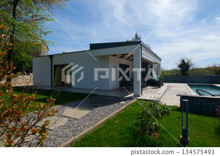 Covered terrace of a modern modular house with dining table, overlooking a swimming pool and gabion fence. Contemporary prefabricated home architecture with outdoor living space 134575491