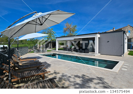 Modern modular house backyard featuring a rectangular swimming pool, large patio with wooden sun loungers, and umbrellas against a blue sky. Contemporary prefabricated home design Modern modular house backyard featuring a rectangular swimming pool, large patio with wooden sun loungers, and umbrellas against a blue sky. Contemporary prefabricated home design 134575494