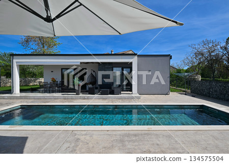 Modern modular house backyard featuring a rectangular swimming pool, large patio with wooden sun loungers, and umbrellas against a blue sky. Contemporary prefabricated home design 134575504