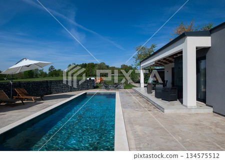 Modern modular house backyard featuring a rectangular swimming pool, large patio with wooden sun loungers, and umbrellas against a blue sky. Contemporary prefabricated home design Modern modular house backyard featuring a rectangular swimming pool, large patio with wooden sun loungers, and umbrellas against a blue sky. Contemporary prefabricated home design 134575512