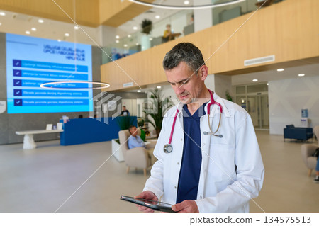 Doctor reviewing patient data on a tablet in a modern hospital lobby while colleagues attend to other tasks 134575513