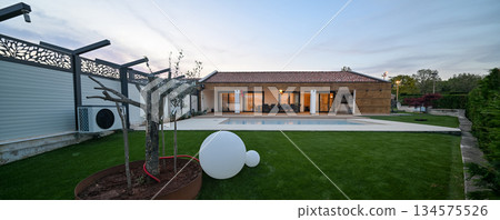 Luxurious modern villa at twilight, featuring a serene swimming pool reflecting warm interior lights. An inviting wooden deck hosts an outdoor dining area, complemented by a stylish hanging chair 134575526