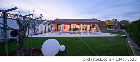 Luxurious modern villa at twilight, featuring a serene swimming pool reflecting warm interior lights. An inviting wooden deck hosts an outdoor dining area, complemented by a stylish hanging chair 134575528