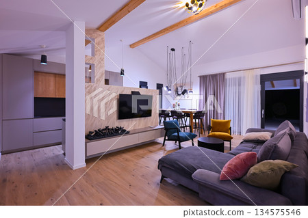 Modern open-plan living and dining area with an integrated kitchen, featuring a comfortable sofa, stylish armchairs, and a bar-height dining table. Wooden floors and ceiling beams enhance this 134575546
